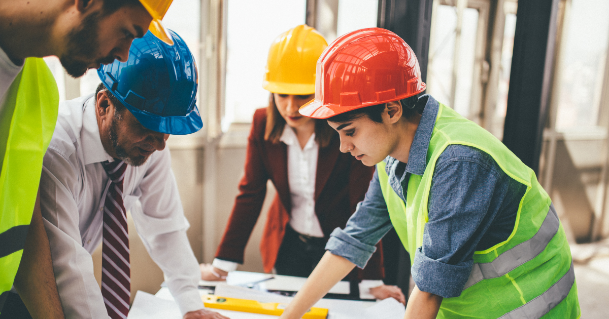 The image shows four construction workers gathered around a table, examining plans or documents. They are indoors with large windows in the background, letting in ample natural light. All four individuals are wearing safety helmets and appear focused on the task at hand. The person on the left, wearing a white hard hat and safety vest, is looking down at the plans. The person in the middle wears a yellow hard hat, with their attention directed at the documents. A person to the right is wearing a red hard hat and a bright green safety vest, closely examining the documents with a forward lean. The fourth person in the background, wearing a blue hard hat, is also engaged in the discussion.